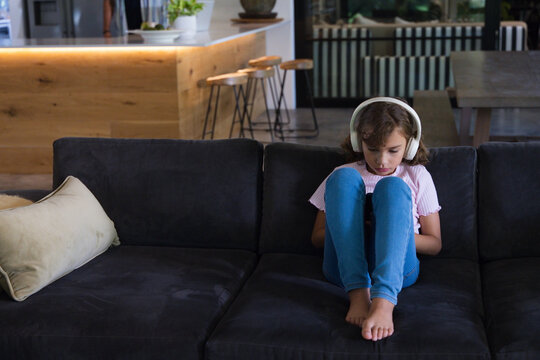 Female child sitting right on grey sofa wearing white over-ear headphones using tablet, copy space