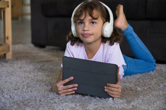 Female child lying on gray carpet at home wearing white headphones and pink top, holding tablet