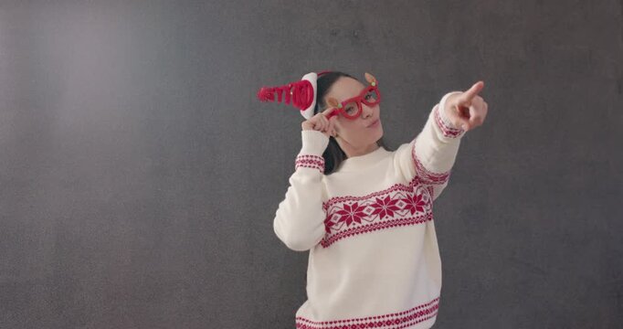 Woman placing red glasses, elf hat, Nordic sweater, posing for fun by grey wall, pointing right