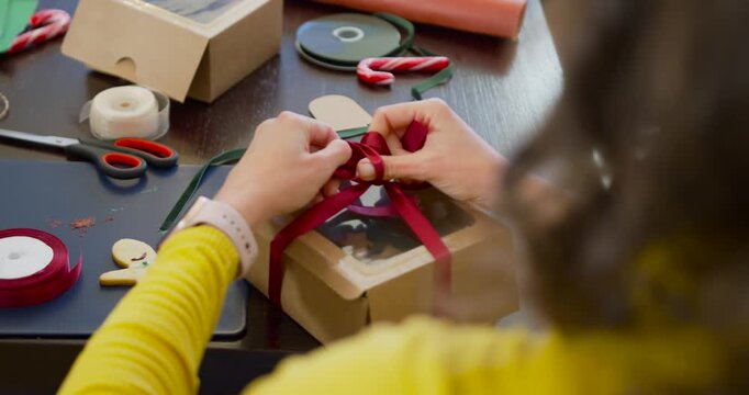 Woman pulling and arranging red ribbon, tying bow to finish windowed gift box in yellow top