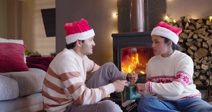 African American couple in Santa hats by woodstove, man handing gift, woman untying bow for holiday