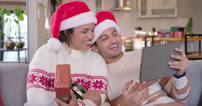 African couple wearing Santa hats sharing holiday tablet call, holding green-ribbon gift on couch