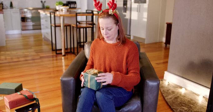 Mature woman in red sweater picking green gift at home, trying to untie ribbon after sound