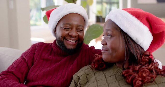 Couple sitting on couch in home living room wearing Santa hats, man touching neck, causing laughter
