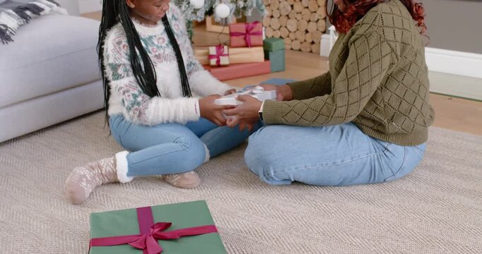 African American mother and daughter sharing white box as child offering gift, hugging by Xmas tree