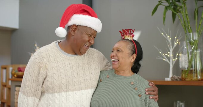 Couple smiling, man placing arm around senior woman, leaning on shoulder at home wearing Santa hat