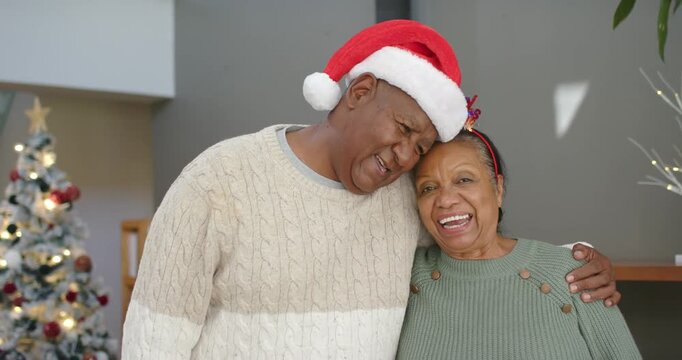 African American couple posing at home, man wearing Santa hat wrapping arm, leaning, woman smiling