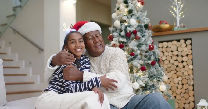 Senior African American grandpa hugging grandchild on couch for holiday session by tree Santa hat