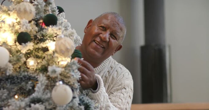 Senior African American man in sweater peeking leaning at tree adjusting decor for holiday at home