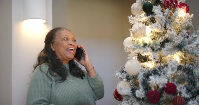 Senior woman taking smartphone call in green sweater, adjusting snowy tree ornament at home