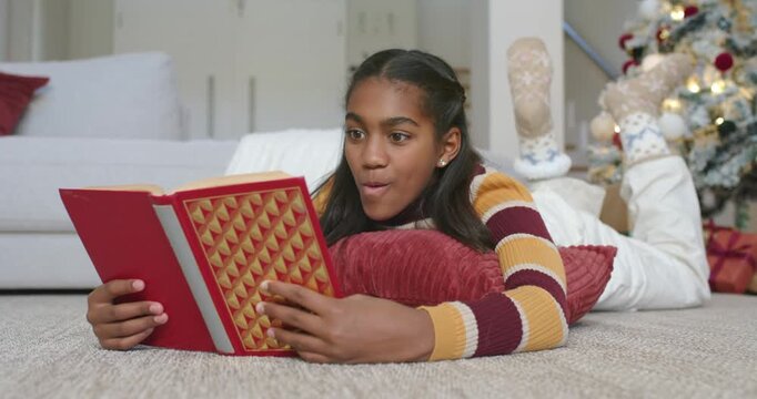 Teen African girl opening red book on cushion, reacting then smiling while reading near Xmas tree