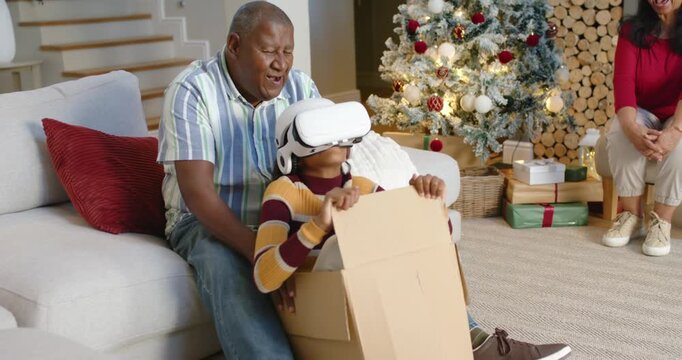 African American family watching youth woman wearing VR in box at home, man holding, senior smiling