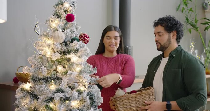 African American couple decorating tree for Xmas at home she taking ornament from basket hanging