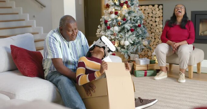 Family member wearing white VR headset testing holiday gift in cardboard box while man steadying