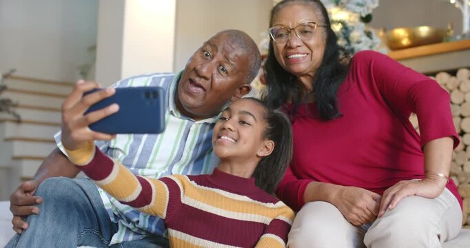 African American family teen girl holding phone on couch, framing selfie while seniors lean smiling