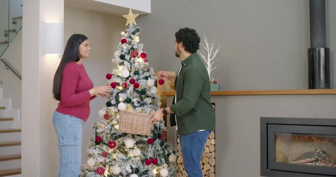 Couple decorating Christmas tree at home selecting ornaments from wicker basket and hanging baubles