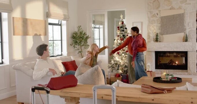 Diverse friends entering doorway with luggage, hugging, reuniting in living room by Christmas tree