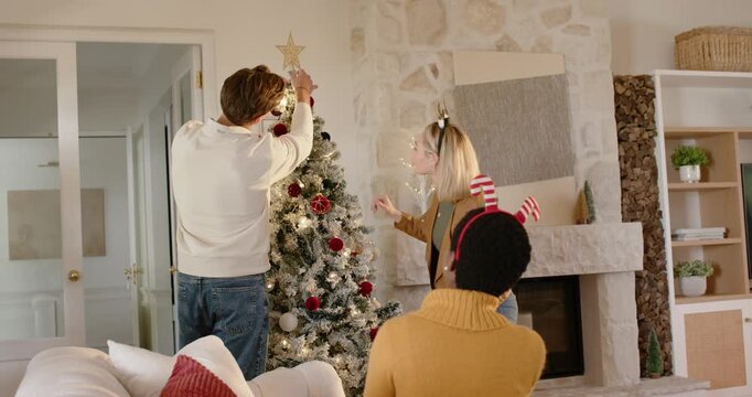 Diverse trio trimming tree at home, tall man placing star, blonde with striped band, tuning lights