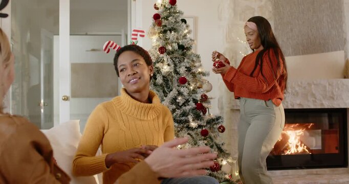 African American women inspecting ornament, placing red baubles on tree while chatting for holidays