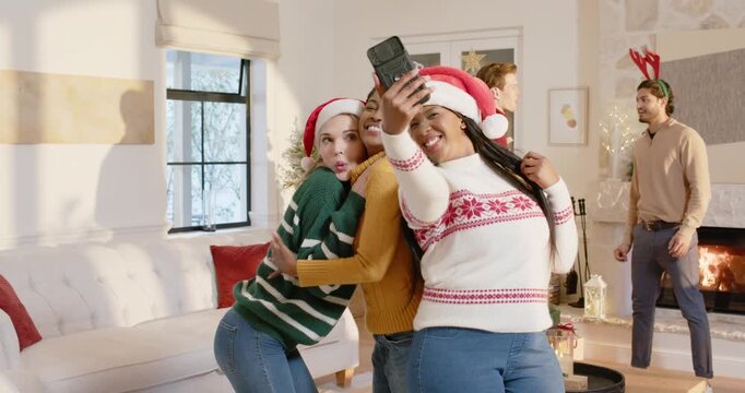 Diverse female friends in Santa hats posing selfie at home celebrating as right woman raising phone