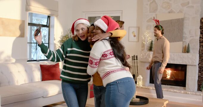 Diverse women friends posing at home, left woman extending smartphone, capturing holiday memory