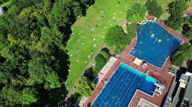 Aerial view of public outdoor swimming pool in Berlin city park with people sunbathing on green lawn during summer.