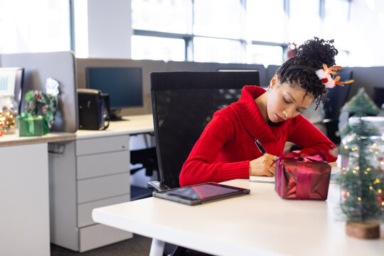 African American woman writing on notepad at office in red sweater with tree, gift, copy space