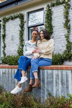 Diverse female friends sitting on ledge at brick wall holding hands wearing Christmas headbands