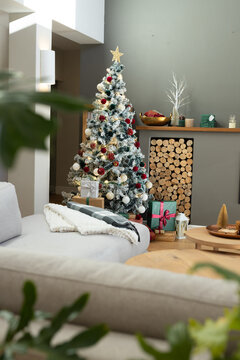 Flocked Christmas tree is standing in modern living room with wrapped gifts, lit mantel