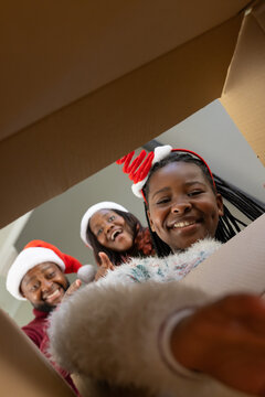 African American family wearing Santa hats leaning over box at home opening gift with ornament