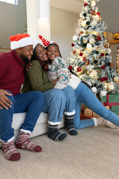African American family sitting on sofa hugging, in sweaters, Santa hat beside lit Christmas tree
