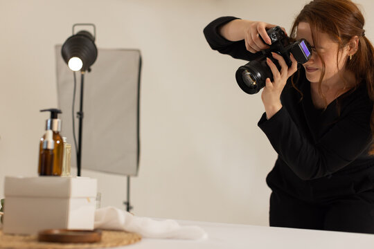 Female photographer in dark top leaning, aiming DSLR at table with boxes, softbox, copy space