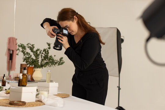 Female photographer leaning over tabletop in studio holding DSLR, shooting skincare with light