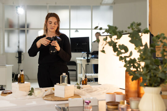 Diverse coworkers female photographer wearing black adjusting DSLR on table with scrim and bottles