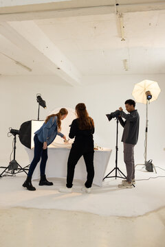 Diverse photography team in studio, photographer operating tripod camera, stylists arranging props