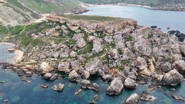 rocky limestone headland dividing two turquoise bays boulder field hiking trail clear shallow water sandy beach coves either side malta west coast aerial drone footage
