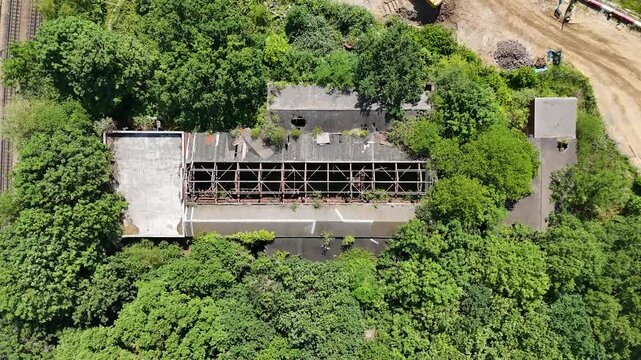 Top down view of a derelict railway building in west London with a collapsed roof and rusting steel frame, overgrown by trees beside a bare earth haul road