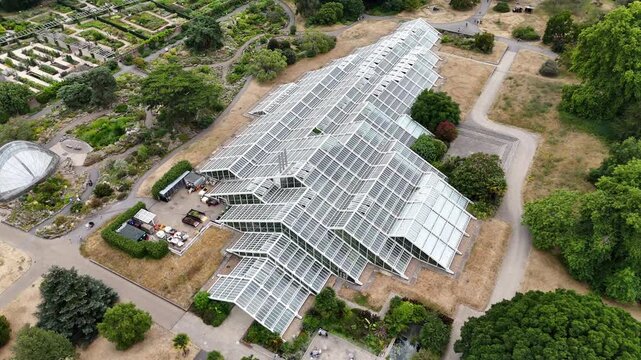 Angular glass roofs of a modern conservatory step across the ground in botanic gardens, surrounded by dry brown lawns