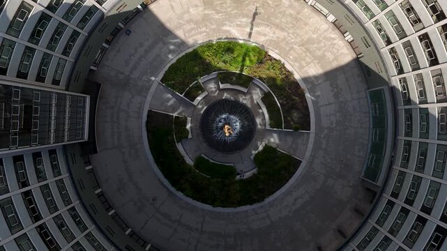 Looking straight down into the paved circular courtyard and fountain at the centre of a ring shaped modernist building
