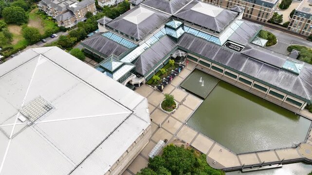 Angular grey roofs and glazed ridges of a large modernist building step down to an ornamental pond and terrace