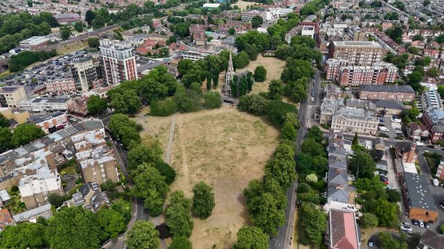 Victorian church stands on a large common where the grass has been burnt brown by weeks of summer heat, ringed by shops and flats