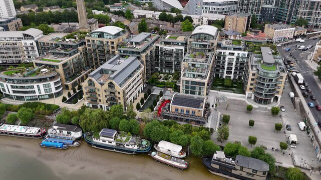 Houseboats and barges moored along the riverbank beside a modern development of brick and glass apartments