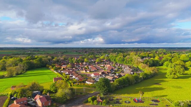 Aerial view shows a peaceful village surrounded by lush green fields. Red-tiled homes are scattered across the landscape. Dense trees and open grasslands frame the settlement