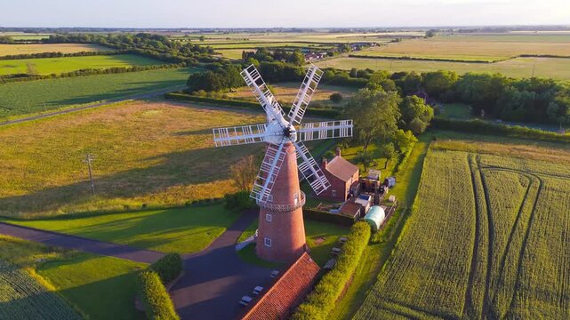 Red windmill stands tall in green fields. Sunlight glows softly on historic structure. Surrounding farmland stretches to horizon. Calm rural setting evokes peace and tradition. Lincolnshire