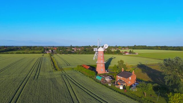 Aerial view of windmill beside green fields under clear blue sky. Windmill stands tall amid vibrant farmland with neat crop rows. Sunlight glows bright. Perfect for agricultural, rural.