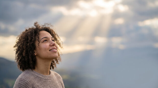 Young woman looking up at sunbeams breaking through clouds with a hopeful expression