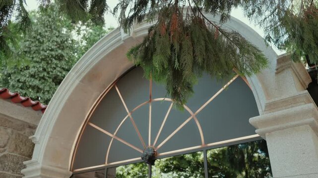 Arched fanlight window in a stone facade framed by hanging pine branches and ivy