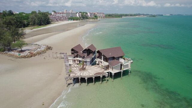 Drone descends toward a derelict seaside villa built above turquoise water, revealing aging hospitality infrastructure and an empty beachfront setting along the Malaysian coast.