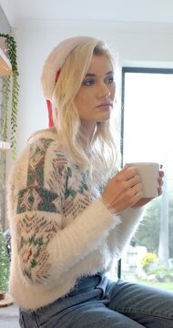 Vertical video: Woman at counter holding mug and sipping warmth in Santa hat, gazing out window