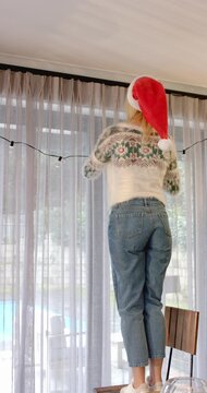 Vertical video: Lifting lights, woman is climbing chair and draping bulbs on curtain rod for decor